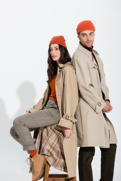 stylish woman in trench coat sitting on stool and looking away near trendy man on white - Foto, Bild