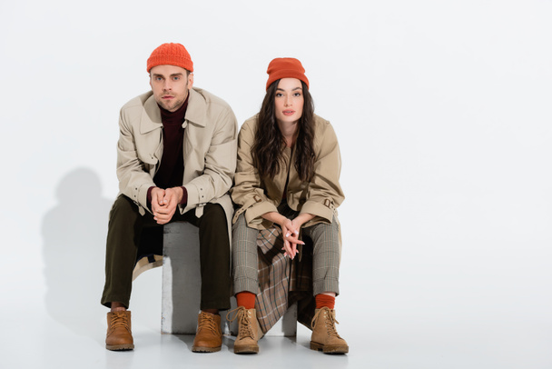 young brunette woman in trench coat sitting with clenched hands near trendy man in beanie hat on white - Foto, Bild