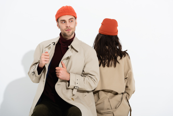 trendy man in beanie hat touching trench coat near woman sitting on white - Photo, Image