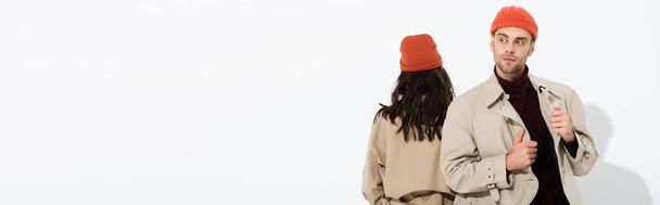 πανοραμική φωτογραφία του κομψό άνδρα σε καπέλο beanie αγγίζοντας καμπαρντίνα κοντά γυναίκα απομονωμένη σε λευκό - Φωτογραφία, εικόνα