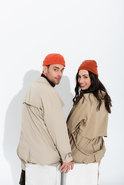 trendy paar in beanie hoeden en trench jassen zitten en kijken naar camera op wit  - Foto, afbeelding