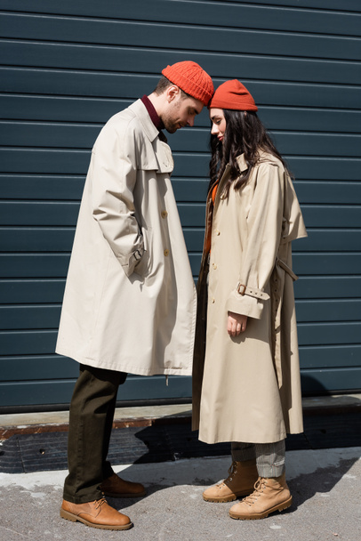 full length of stylish couple in trench coats and hats leaning on each other outside - Φωτογραφία, εικόνα