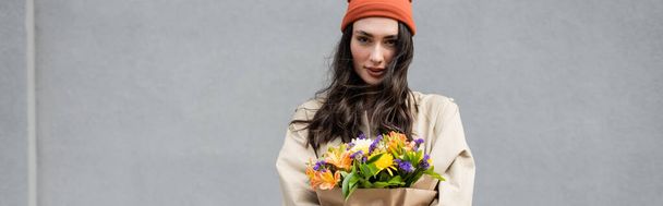 panoramic shot of stylish woman in beanie hat and trench coat holding flowers near grey wall - Photo, Image