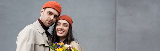 panoramic crop of trendy couple in trench coats and hats near bouquet of flowers  - Foto, Imagem