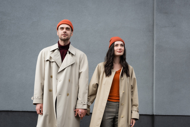 elegante pareja en abrigos de trinchera y sombreros de pie y tomados de la mano cerca de la pared gris exterior - Foto, Imagen