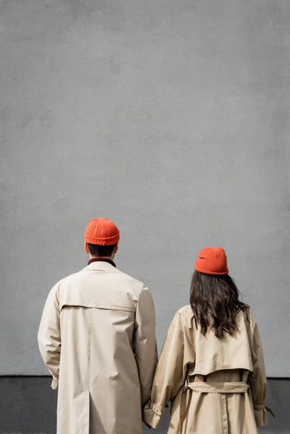vista posteriore di coppia in trench e cappelli in piedi vicino alla parete grigia - Foto, immagini