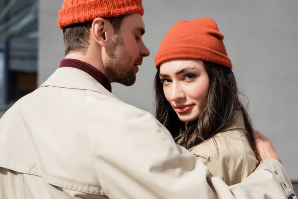 κομψό άνδρα σε καμπαρντίνα παλτό αγκαλιάζει γυναίκα σε καπέλο beanie έξω - Φωτογραφία, εικόνα