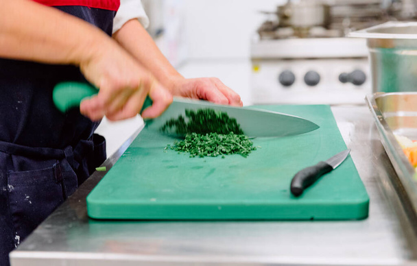 Mulher velha não reconhecida cortando salsa fresca em uma cozinha de residência sobre uma mesa verde com duas facas diferentes - Foto, Imagem