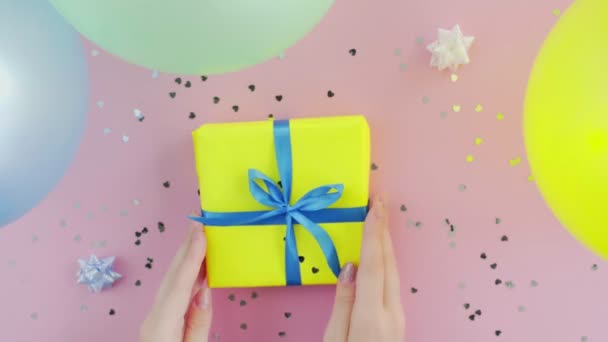 Žena se dotkne vánoční dárek box zabalený ve žlutém papíru s modrým lukem na růžovém stole - Záběry, video