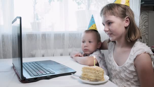 kinderen zitten aan tafel met laptop computer, blazen aangestoken kaars op verjaardagstaart - Video