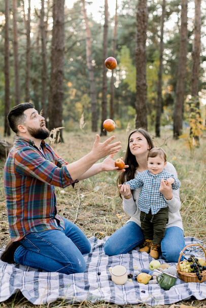 Family, fun and leisure concept. Portrait of happy family sitting on checkered plaid with fruits in autumn forest. Handsome father, juggling apples, while his wife and little son having fun - Fotó, kép