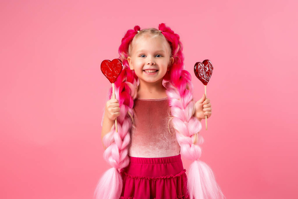 Malá šťastná holčička s velkými růžovými copánky baví s lízátkem ve tvaru srdce. Obrázek s prostorem pro text izolovaný na růžovém pozadí, koncept dětských emocí. - Fotografie, Obrázek
