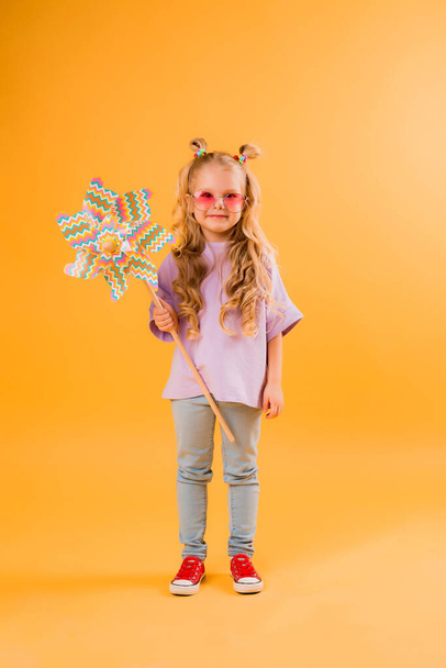 Menina alegre bonito pouco em grandes óculos de sol rosa e pinwheel brincando. Isolado em fundo laranja, conceito de emoções infantis. - Foto, Imagem