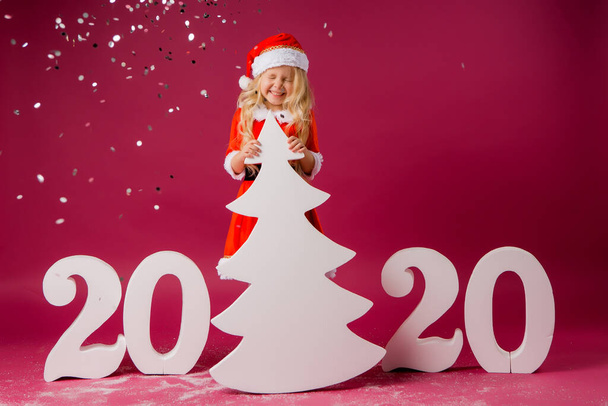 Niña rubia en un traje de Santa sobre fondo rojo con grandes números 2020 y árbol de Navidad. Celebración de la Navidad y el concepto de año nuevo - Foto, Imagen