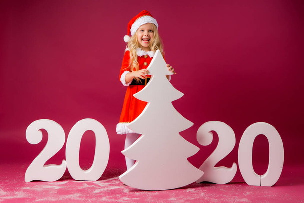 Kleines blondes Mädchen im Weihnachtsmannkostüm auf rotem Grund mit großen Zahlen und Weihnachtsbaum. Weihnachtsfeier und Neujahrskonzept - Foto, Bild