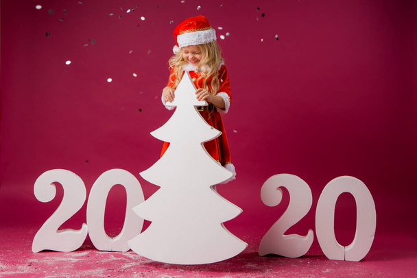 Malá blondýnka v kostýmu Santa na červeném pozadí s velkými čísly roku 2020 a vánočním stromečkem. Oslava vánoční a novoroční koncepce - Fotografie, Obrázek