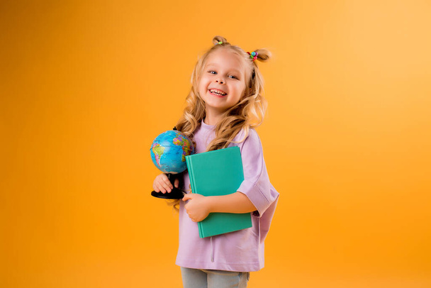 Wesoła blondynka trzymająca kulę i księgę w rękach. Odizolowane na pomarańczowym tle, dziecięce emocje i powrót do koncepcji szkoły.  - Zdjęcie, obraz