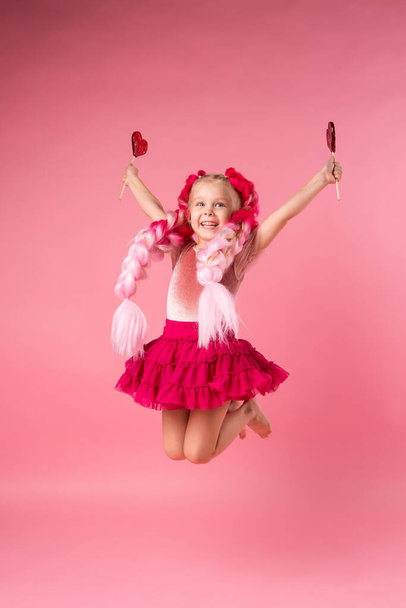 Klein gelukkig meisje met grote roze vlechten die plezier hebben met hartvormige lolly 's. Beeld met ruimte voor tekst geïsoleerd op roze achtergrond, emotioneel concept voor kinderen. - Foto, afbeelding