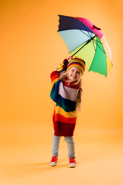 Фото маленькой девочки в огромном свитере и вязаной шляпе с зонтиком, изолированным на оранжевом фоне с пробелами для текста - Фото, изображение