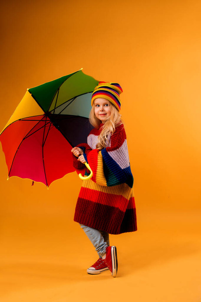 Photo of a little girl in oversize sweater and knitted hat holding an umbrella isolated on orange background with a space for text - Fotó, kép