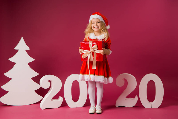 Little blond girl in a Santa's costume with big gift box on red background with 2020 big numbers. Celebration of Christmas and new year concept - Фото, зображення