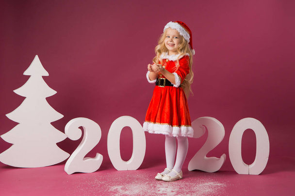 Cute little blond girl in a Santa's costume on red background with 2020 big numbers. Celebration of Christmas and new year concept - Foto, immagini