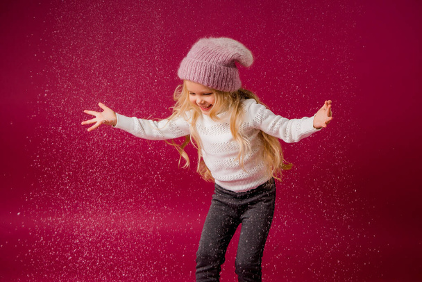 Μικρό χαρούμενο κορίτσι με ζεστά χειμωνιάτικα ρούχα διασκεδάζει στο κόκκινο φόντο. Αντιγραφή εικόνας χώρου με τοποθέτηση για το κείμενο ή τη διαφήμισή σας - Φωτογραφία, εικόνα