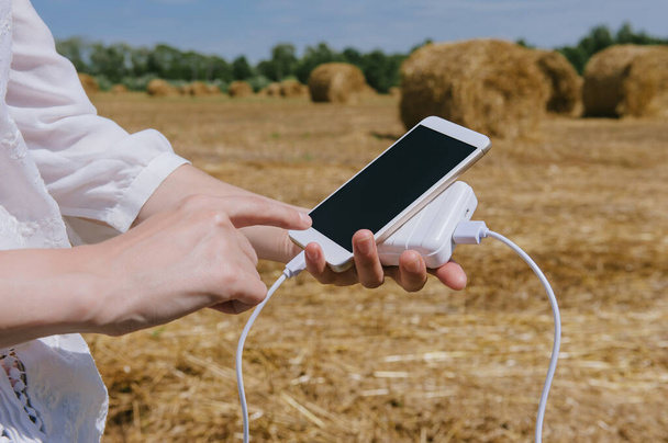 Mock Up of a smartphone and the Power Bank, in the hand of a girl. against the background of a field with haystacks. - Фото, изображение
