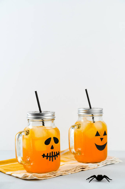 Halloween pumpkin mocktails in glass jars decorated with scary faces on the gray table, white background. - Foto, afbeelding