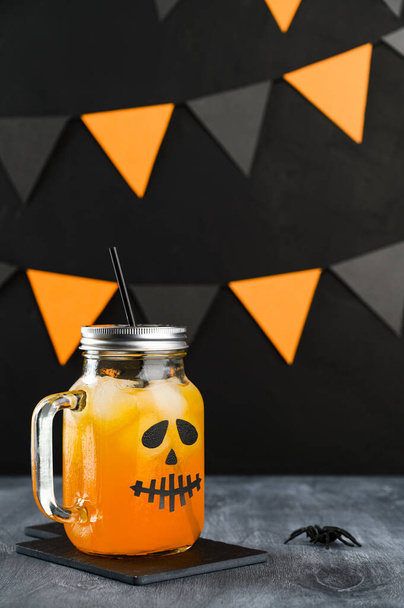 Halloween pumpkin iced mocktail in glass jar decorated with scary face on a dark table. DIY Halloween Party decoration on black wall. - Foto, Imagem