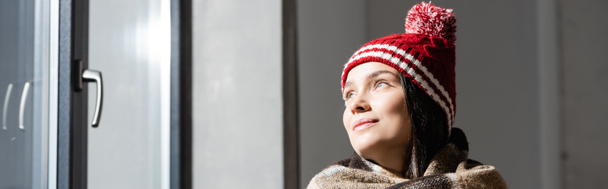 ιστοσελίδα κεφαλίδα της νεαρής γυναίκας σε ζεστό πλεκτό καπέλο κοιτάζοντας μακριά, ενώ στέκεται κοντά στο παράθυρο στο σπίτι - Φωτογραφία, εικόνα