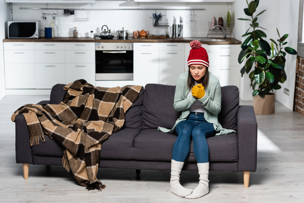 zitternde Frau mit warmer Mütze, Handschuhen und Socken auf Sofa neben karierter Decke in der Küche - Foto, Bild