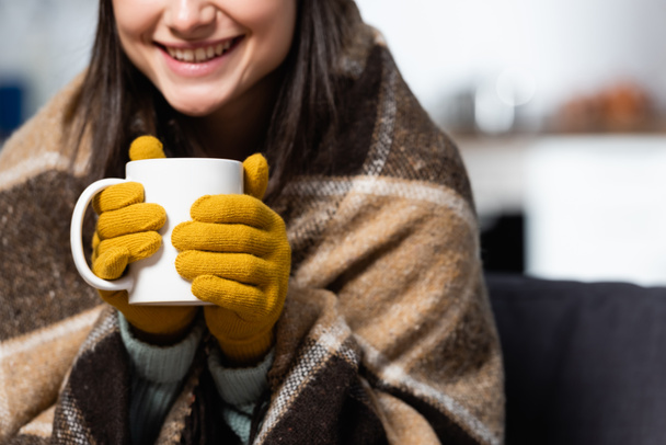abgeschnittene Ansicht einer Frau mit Strickhandschuhen, eingewickelt in eine karierte Decke, die zu Hause eine Tasse warmen Tee hält - Foto, Bild