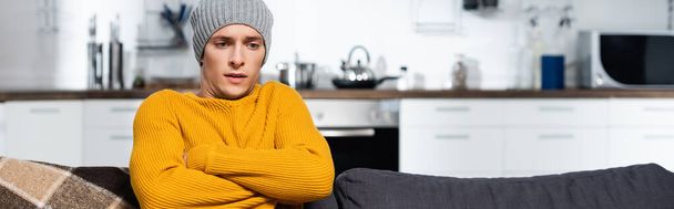 horizontal crop of freezing man in warm sweater and hat sitting with crossed arms in cold kitchen - Foto, Imagen