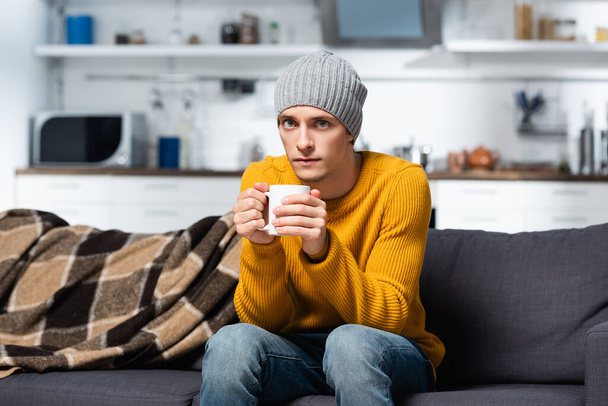 hombre frío en suéter de punto y sombrero mirando a la cámara mientras sostiene la taza de té caliente en la cocina - Foto, imagen