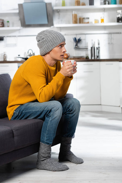 freezing man in knitted sweater, hat and socks holding cup of warm beverage in kitchen - Fotografie, Obrázek