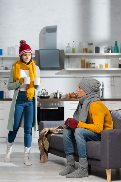 femme en vêtements chauds portant des tasses de boisson chaude tandis que petit ami assis sur le canapé dans la cuisine froide - Photo, image