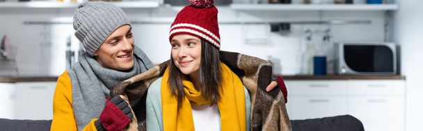 horizontal image of man in knitted hat wrapping joyful girlfriend with warm plaid blanket while sitting in cold kitchen - Photo, image