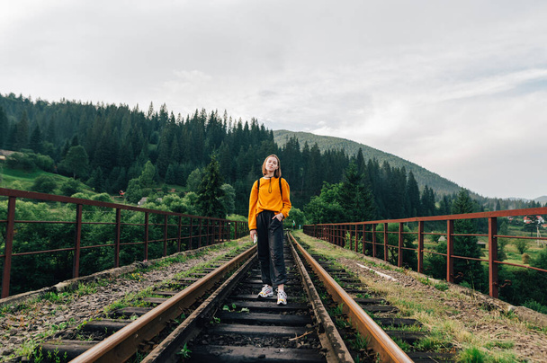 Позитивная женщина в стильной повседневной одежде стоит на мосту с железнодорожной дорожкой на фоне горного пейзажа, глядя в камеру с улыбкой на лице. Ворохта, Карпаты - Фото, изображение