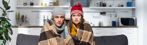 Ein frierendes Paar mit warmen Hüten, das in der kalten Küche unter einer karierten Decke in die Kamera blickt - Foto, Bild
