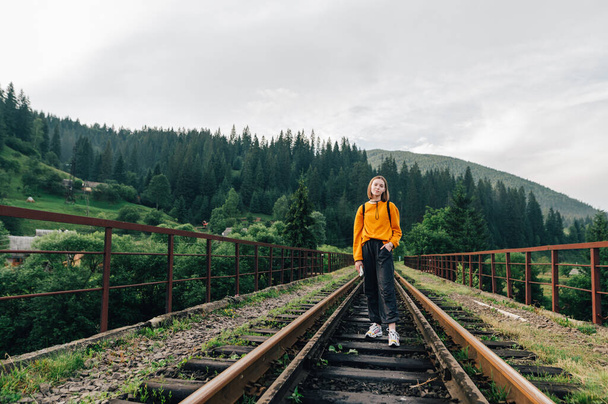 Стильная пешеходная женщина стоит на железнодорожной трассе в горах и позирует перед камерой. Портрет в полный рост. Hipster travels.Vorokhta, Карпаты, Украина - Фото, изображение