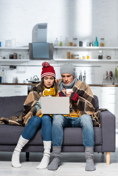 cold couple in warm hats and gloves sitting on sofa under plaid blanket and watching movie on laptop - Foto, Imagem