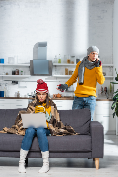 mujer congelada sentada en el sofá con el ordenador portátil mientras su novio preocupado hablando en el teléfono inteligente en la cocina fría - Foto, Imagen