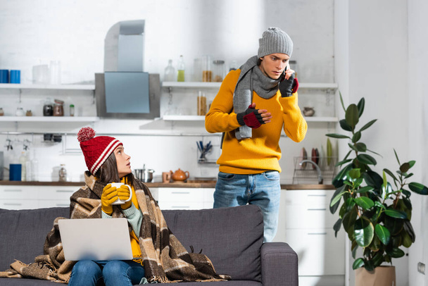 kalte Frau mit warmer Mütze und Handschuhen auf Sofa unter karierter Decke sitzend, während besorgter Freund am Smartphone spricht - Foto, Bild