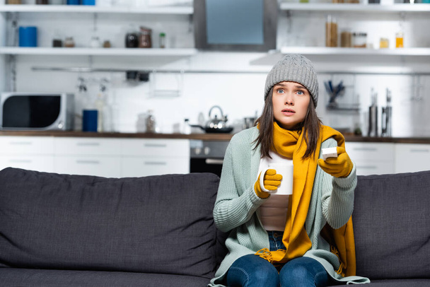 frozen woman in knitted clothes holding tv remote controller and cup of warming beverage while sitting on sofa in cold kitchen - Photo, Image