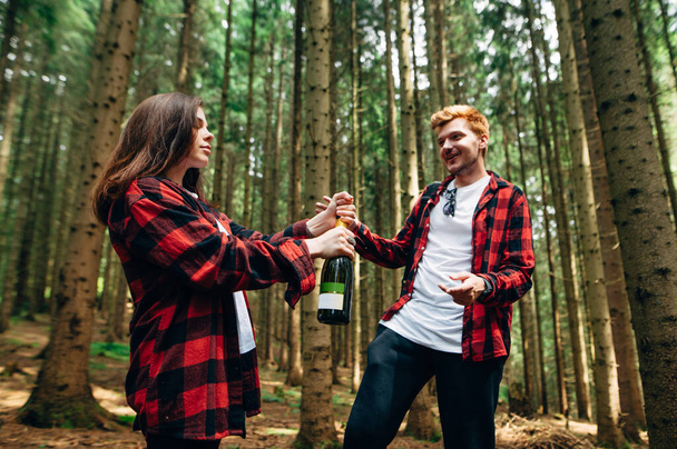 Pareja feliz en camisas rojas abriendo champán en el bosque en un paseo. Mujer arranca una botella de vino en el bosque en un paseo con su novio. - Foto, imagen