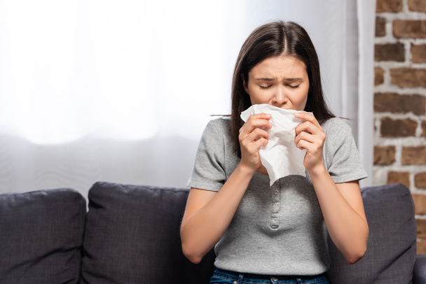 ill woman sneezing in paper napkin while sitting on sofa at home - Fotó, kép