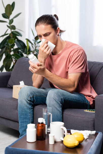 uomo malato pulire il naso con tovagliolo di carta mentre seduto sul divano e in possesso di bottiglia con pillole - Foto, immagini