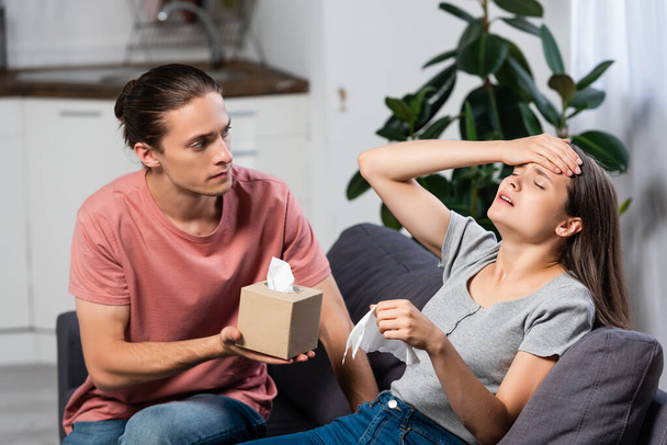 young man holding paper napkins near ill girlfriend suffering from migraine with closed eyes - Photo, Image
