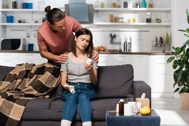 Mann berührt kranke Frau auf Sofa in Küche und hält Flasche mit Tabletten - Foto, Bild